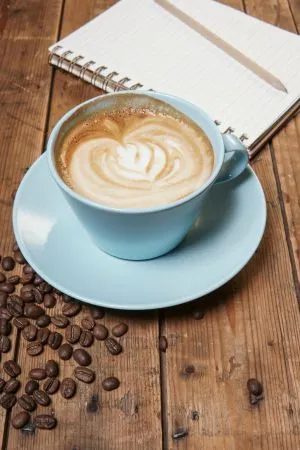 Photo d'une tasse à café bleu ciel sur une table en bois , sur laquelle sont posés des grains de café et un cahier de brouillon pour illustrer un rendez vous de travail